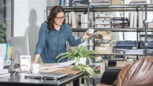 Businesswoman reviewing documents in an office with shelves, files, and workspace items.